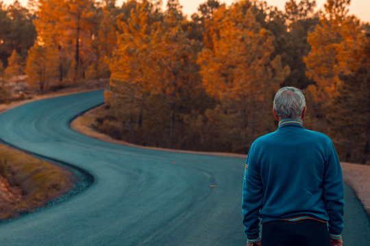 Active Senior Man Of Back Walking On Lonely Highway For The Mountains