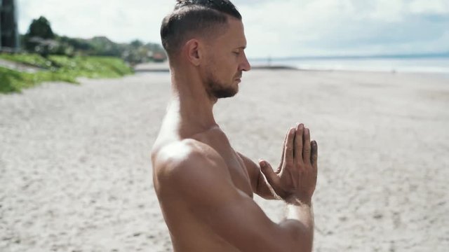 Close-up slow motion steadicam shot, athletic man doing yoga on beach with his hands together and eyes closed