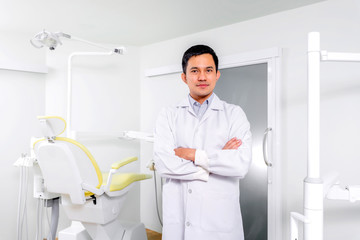 Cheerful Asian dentist standing in his clinic with his dental chair