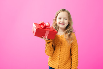 Happy little child girl in a warm knitted sweater holding a box with a gift, on pink background....