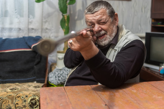 Bearded Caucasian Senior Aiming His Walking Stick Like A Gun Sitting At The Table Indoor