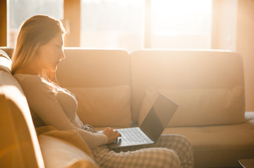 Beautiful girl looking into the laptop screen, sitting on the couch. Beautiful morning light fills the room in the winter morning.