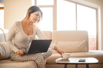 Fototapeta premium Attractive blond girl in home clothes stretches for a cup of tea, looking up from work at a laptop