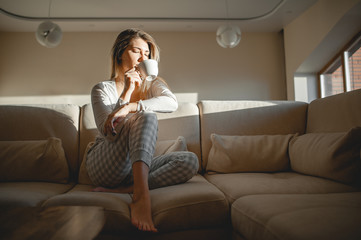 Obraz premium A blonde girl after breakfast sits in her pajamas in a large room on a sofa and drinks tea or coffee from a mug.