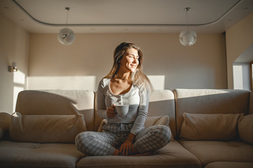 Obraz premium Beautiful girl in pajamas sitting in the lotus position with a mug of warm tea in her hands and looking with a smile at the morning light