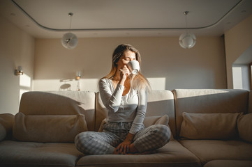 A blonde girl after breakfast sits in her pajamas in a large room on a sofa and drinks tea or coffee from a mug.