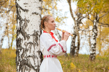 Beautiful girl with braids in Slavic clothes playing the flute in the forest