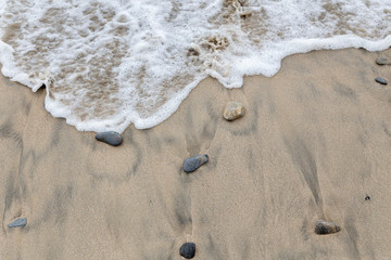 Rough water on the sand. On the surface of the water there are small waves and foam. On the sand are stones. There are dark patches in the sand. Sunny weather. Ocean or sea. Empty space.