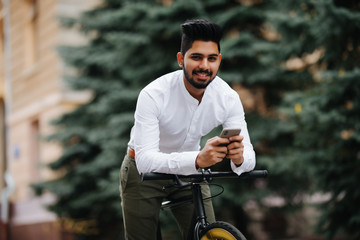 Chilling after good ride. Handsome young indian man holding mobile phone while standing near his bicycle