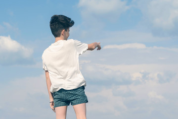 Boy in the T-shirt and shorts shows his finger on the cloudy blue sky. Summer and travel concept