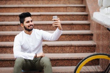 Excited young asian man sitting on stairs with bicycle, taking a selfie with mobile phone