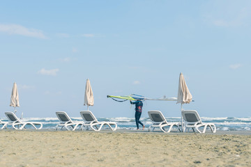 Fototapeta na wymiar Windsurfer with board on the Cyprus beach. Sea view, umbrellas and sunbeds on sunset.