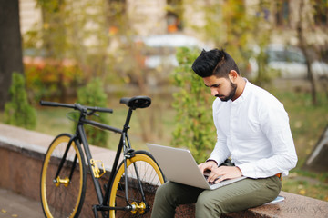 Fototapeta premium Handsome young indian businessman dressed in suit working on laptop computer while sitting outdoors near bicycle