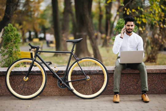 Portrait of young indian cheerful handsome man early morning with bicycle using laptop computer and Talking by phone outdoors