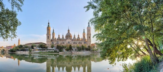 A landscape of christian Pilar Cathedral and Santiago Bridge reflecting in the Ebro river during a sunny summer day in Zaragoza, Aragon region, Spain