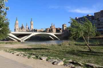 The Pilar Cathedral and the Santiago Bridge seen from the Ebro river promenade with trees and lawn...