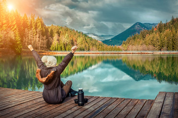 Happy young woman sitting on the wooden pier with hands up, mountains