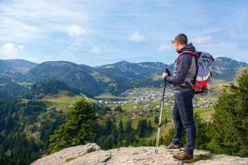 Hiker young man with backpack and trekking poles standing on edge of cliff and looking at the lake, rear view