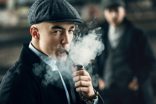 stylish men smoking in retro clothes posing on background of railway. england in 1920s theme. fashionable look of brutal confident man. atmospheric moments with smoke
