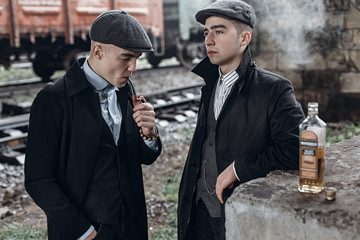 stylish gangsters men, smoking. posing on background of railway with bottle of alcohol. england in 1920s theme. fashionable brutal confident group. atmospheric  moments