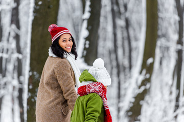 smiling african american mother walking with daughter  and looking at camera in winter park