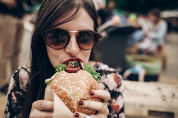 stylish hipster woman eating juicy burger. boho girl biting yummy cheeseburger, smiling at street food festival. summertime. summer vacation travel picnic. space for text