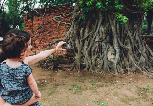 Thailand, Ayutthaya, Mother and daughter taking a photo of the Buddha head in between tree roots at Wat Mahathat