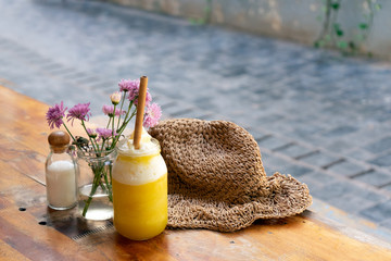A jar of fresh yellow pineapple and mango smoothie with a bamboo straw on the table with a straw hat and a vase of pink flowers near it. Summer concept. No more plastic straw concept