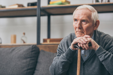 sad retired man with grey hair holding walking cane in living room
