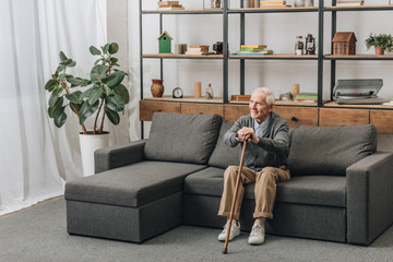happy senior man smiling and holding walking cane while sitting on sofa