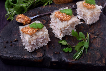 Holodets chicken jelly cut into cubes topped with mustard and cilantro on a dark wooden board surrounded by herbs and pepper