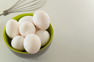 White chicken eggs in a bowl and a whisk on a white background, space for text