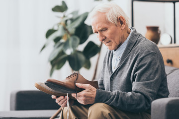 senior man holding shoes in hands while sitting on sofa - obrazy, fototapety, plakaty