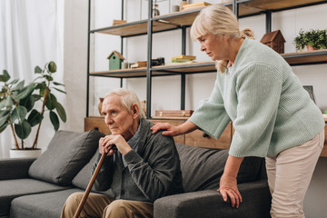 senior wife standing near retired husband sitting on sofa - Powered by Adobe