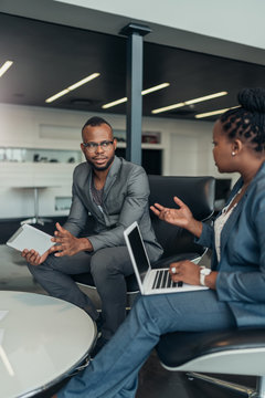 Two African Business People Talking Seriously With Hand Gestures During A Meeting Using Their Laptop And Tablet