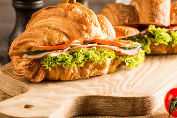 Photo of tasty and fresh croissant sandwich with salad, ham, cheese, tomatoes on wooden background. Morning breakfast concept. Healthy and fast food.