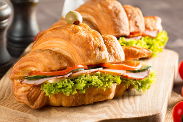 Photo of tasty and fresh croissant sandwich with salad, ham, cheese, tomatoes on wooden background. Morning breakfast concept. Healthy and fast food.
