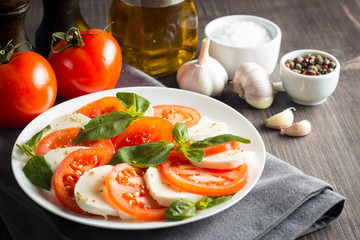 Photo of Caprese Salad with tomatoes, basil, mozzarella, olives and olive oil on wooden background. Italian traditional caprese salad ingredients. Mediterranean, organic and natural food concept.