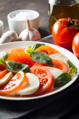 Photo of Caprese Salad with tomatoes, basil, mozzarella, olives and olive oil on wooden background. Italian traditional caprese salad ingredients. Mediterranean, organic and natural food concept.