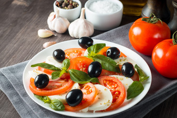 Photo of Caprese Salad with tomatoes, basil, mozzarella, olives and olive oil on wooden background. Italian traditional caprese salad ingredients. Mediterranean, organic and natural food concept.