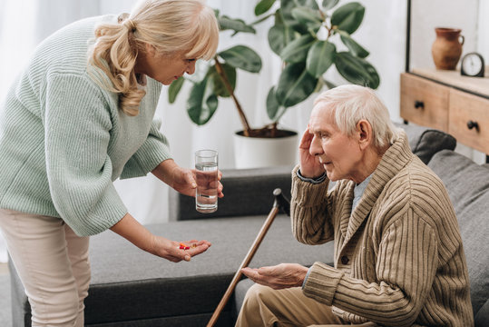 senior woman giving pills to old man with walking stick
