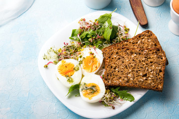 Healthy breakfast: Hard boiled eggs, fresh radish sprouts, arugula and dark whole wheat  bread with herb sauce, on blue background