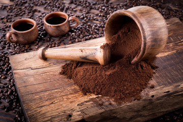 Mortar with ground coffee and pestle lie on a wooden board. Coffee pours from the mortar. Clay mugs stand on coffee beans