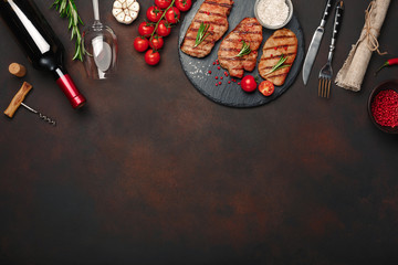 Obraz na płótnie Canvas Grilled pork steaks on stone with bottle of wine, wine glass, knife and fork on rusty background