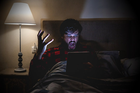 Man in pajamas in bed stirring the tablet or cell phone impregnated at what he sees before sleeping. Touching boy on the verge of sleeping disturbing the scared sleep with what is on the screen in fro