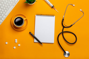 Top view of a doctor's table with notepad and pen stethoscope, keyboard, prescription and pills, a cup of coffee on a yellow background