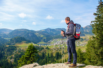 Hiker young man with backpack and trekking poles standing on edge of cliff and looking at the lake, rear view