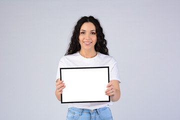 Attractive curly young woman in white t-shirt holding empty blank photo frame with free copy space....