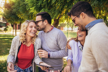 Four happy smiling young friends walking outdoors in the park holding digital tablet