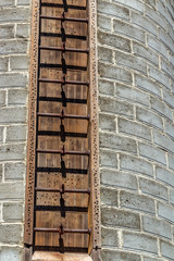 A closeup view of a concrete block silo or windowless tower with a rust metal rung ladder going up the side vertically. The concrete is dark brown to a light gray.